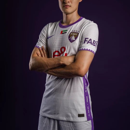 Male soccer player standing with arms crossed, wearing a white and purple kit with team crest and sponsor logos against a dark background.