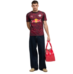 Male model wearing a maroon RB Leipzig-style soccer jersey with Red Bull logo, dark jeans, tan shoes, and holding a red handbag.