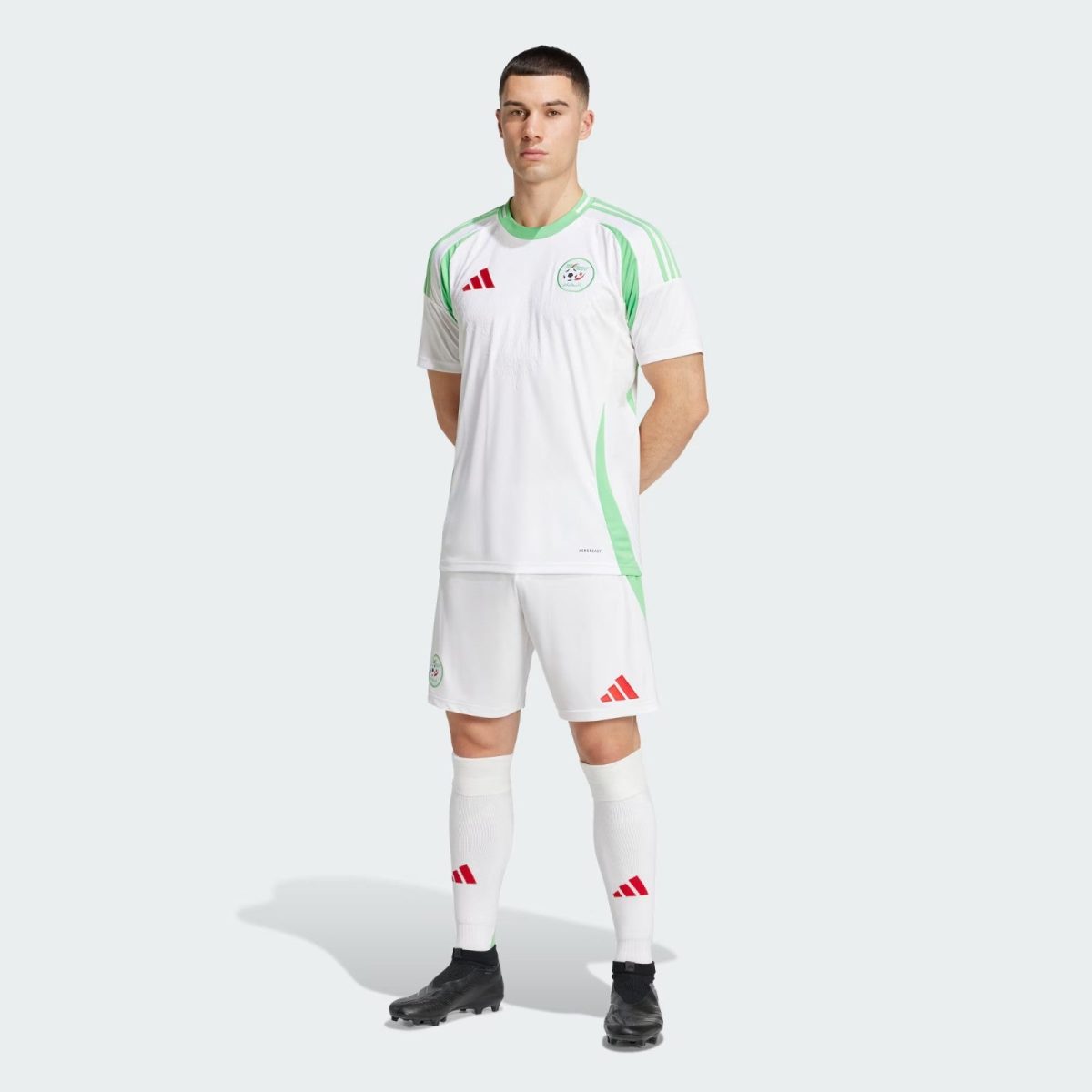 Male soccer player standing with hands behind back, wearing white jersey with green accents and shorts, cleats on a pale background