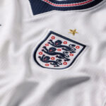 Close-up of a white England national team jersey showing the embroidered crest with three blue lions and red flowers, and a gold star above.