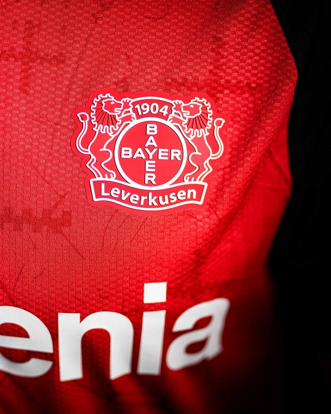 AdJk0KMvYPOK2H5 | UAE SPORT STORE Close-up of a red Bayer Leverkusen football jersey featuring the club crest with '1904' and 'BAYER LEVERKUSEN' text panelled in white.