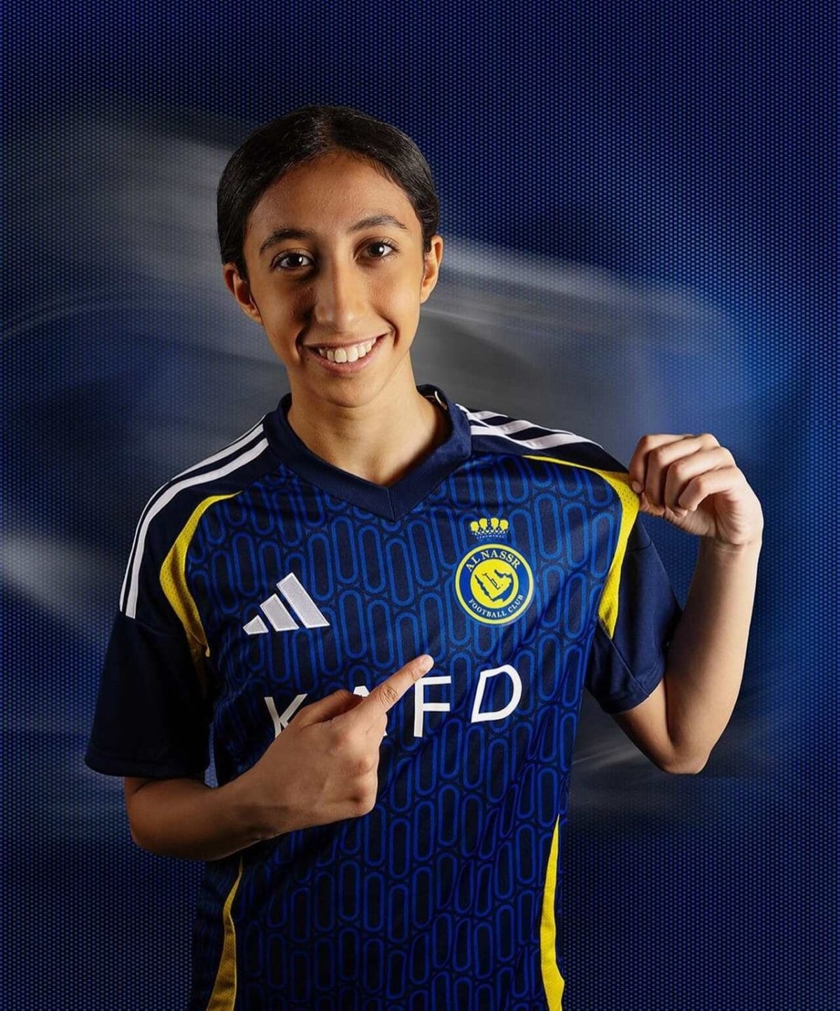 Young person in a blue soccer jersey smiling and pointing to the team crest on the chest against a blue dotted background
