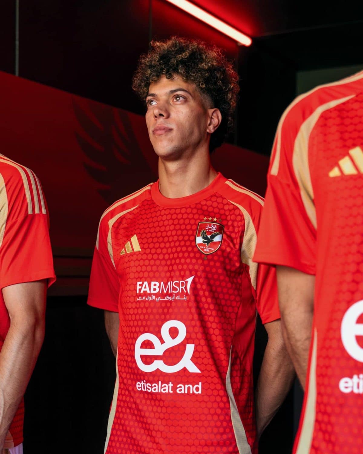 Young soccer player in red jersey looking upward, standing among teammates in a dimly lit room with neon red lighting behind him.