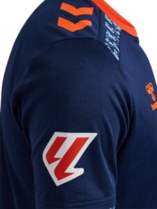 Close-up of a navy blue sports jersey sleeve with orange chevron accents, orange collar trim, and a red-and-white shield logo on the sleeve.