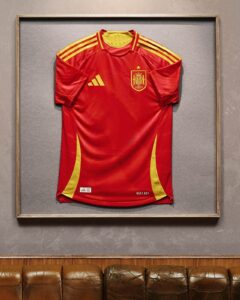 Framed display of a red Spain national football team jersey with yellow accents and crest on a gray wall.