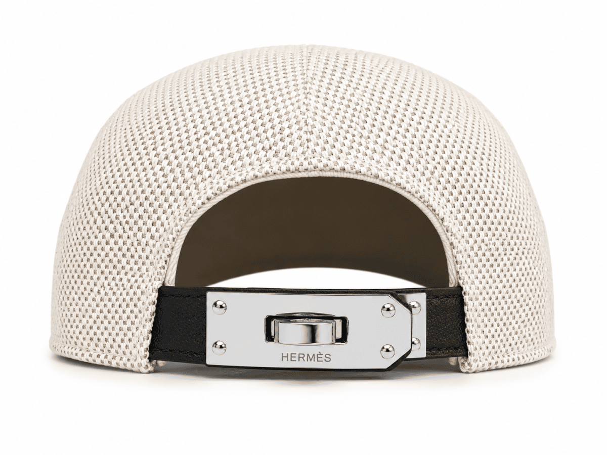 Beige woven cap viewed from the back with an adjustable metal clasp bearing Hermès branding on a black strap.