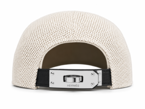 Beige woven cap viewed from the back with an adjustable metal clasp bearing Hermès branding on a black strap.