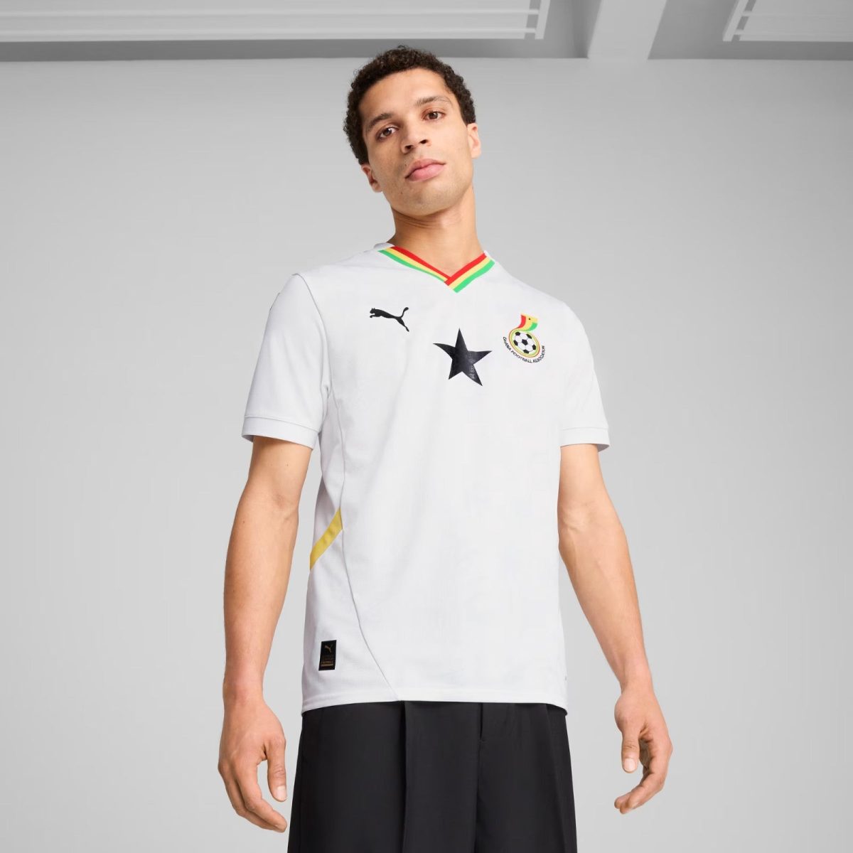 Young man in a white soccer jersey with a black star, Ghana Football Association crest, and Puma logo, standing against a gray background.