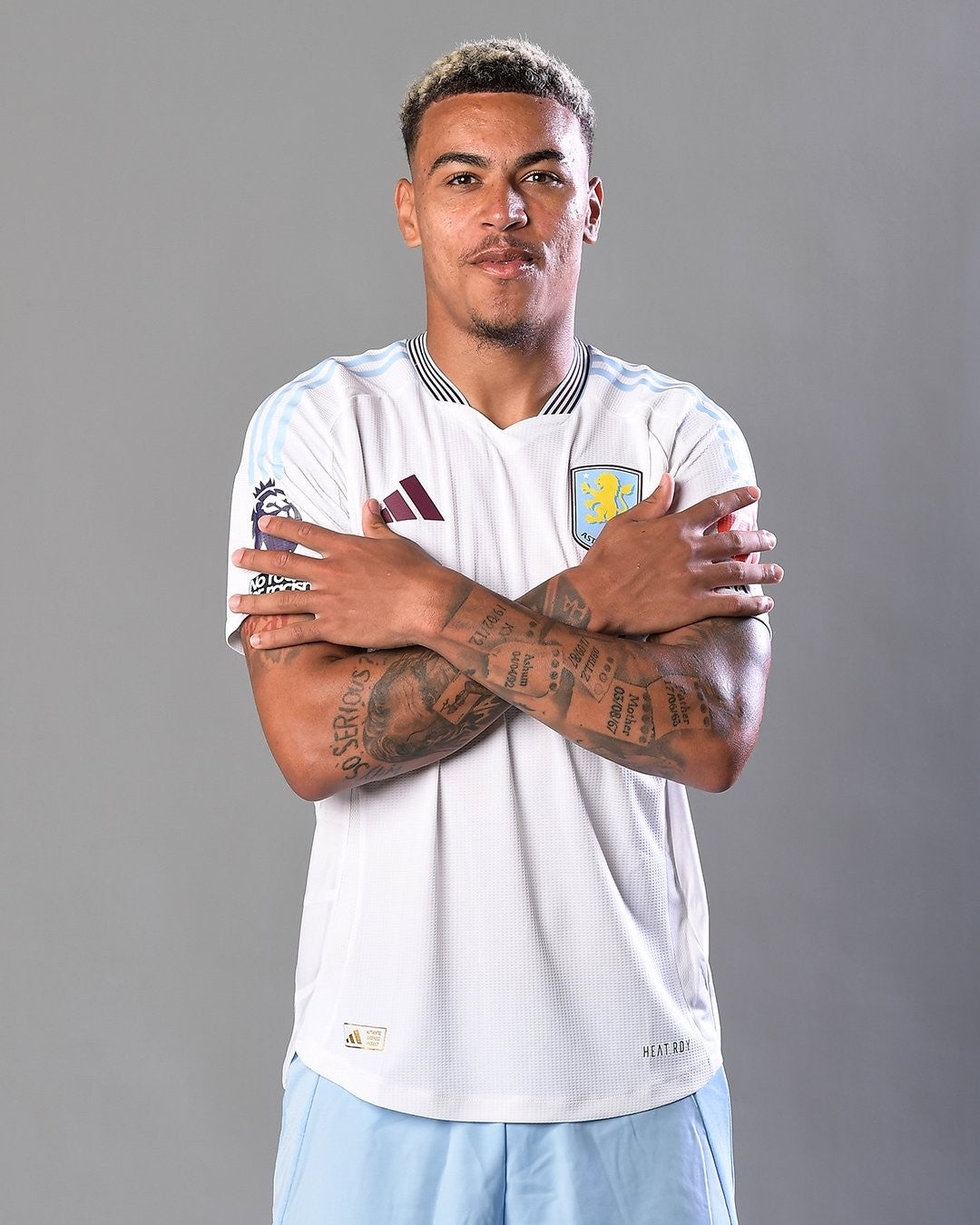 B7ar0c09kB4Ne5Z | UAE SPORT STORE Soccer player in a white jersey and light blue shorts crosses his arms, showing tattoos on both forearms in a studio portrait on a gray background.