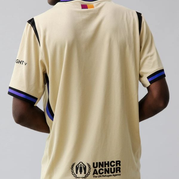 Back view of a beige sports jersey with UNHCR/ACNUR logo and purple-black trim tailing the shoulders and sleeves.