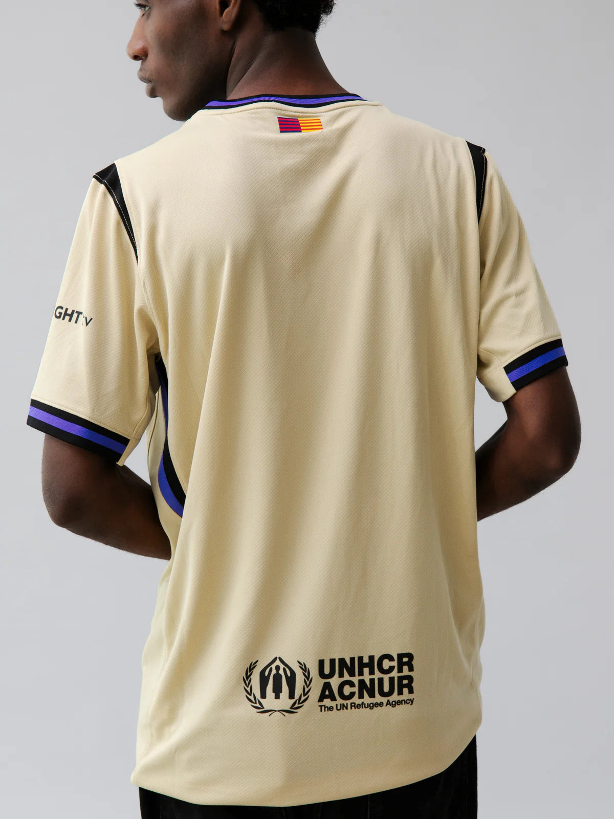 BARCA1-24660 | UAE SPORT STORE Back view of a beige sports jersey with UNHCR/ACNUR logo and purple-black trim tailing the shoulders and sleeves.