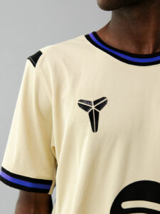 BARCA1-24706 | UAE SPORT STORE Close-up of a cream sports jersey with a black and purple striped collar and a geometric black logo on the chest.