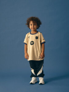 Front-view of a young child standing against a solid blue background, wearing a beige soccer jersey with dark blue trim and white sneakers.