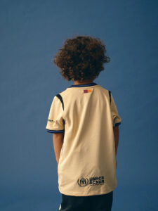 Child with curly hair facing away, wearing a beige UNHCR/ACNUR jersey against a blue background, back view.