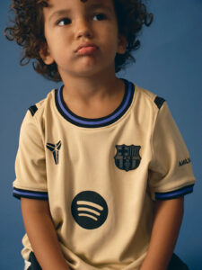 Portrait of a young child with curly hair, looking to the left, wearing a beige FC Barcelona jersey against a blue background.