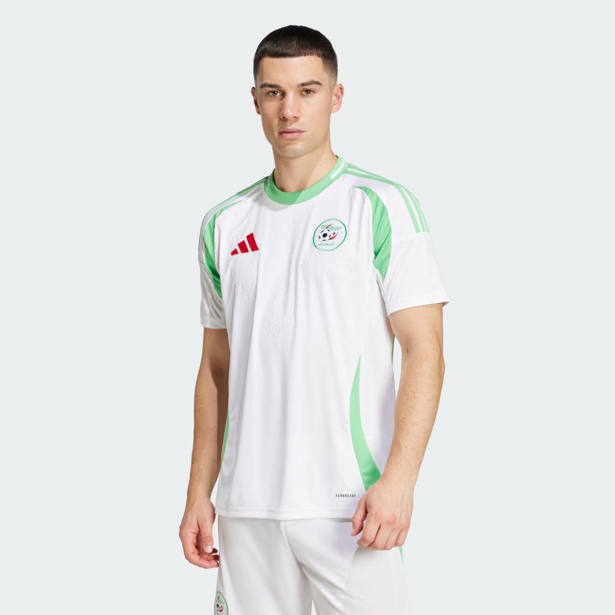 Man in a white soccer jersey with green accents, circular team crest on the left chest, posing against a light gray background.