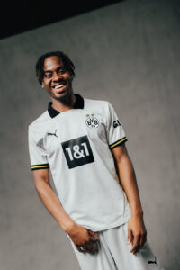 Smiling young man in a white Borussia Dortmund jersey with a black collar, standing against a gray studio backdrop.