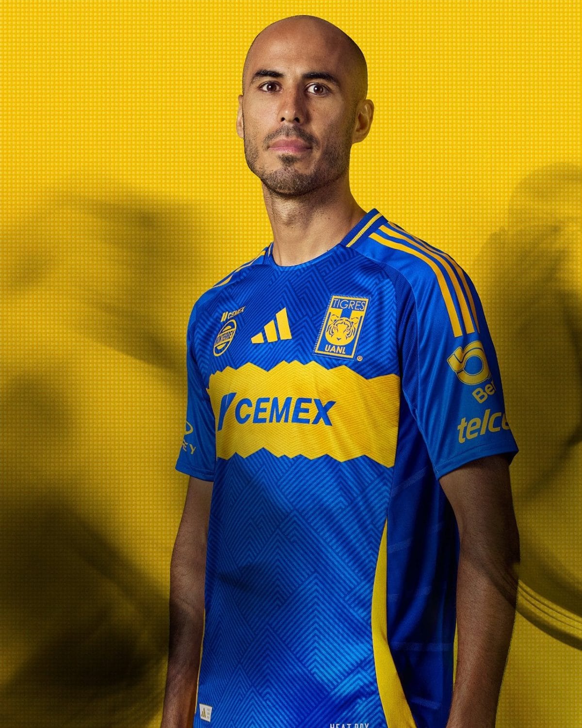 Portrait of a male soccer player wearing a blue and yellow Tigres UANL jersey, standing against a yellow patterned background.”] ,