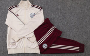 Beige zip-up tracksuit jacket with burgundy stripes worn with matching burgundy pants featuring striped sides and circular crest patches on chest and thigh laid flat on gray carpet
