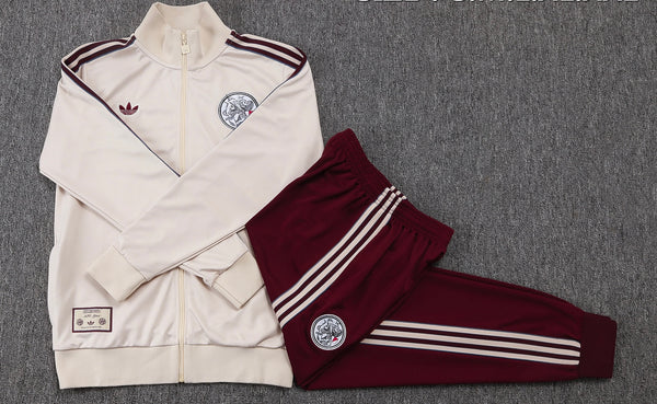 Beige zip-up tracksuit jacket with burgundy stripes worn with matching burgundy pants featuring striped sides and circular crest patches on chest and thigh laid flat on gray carpet