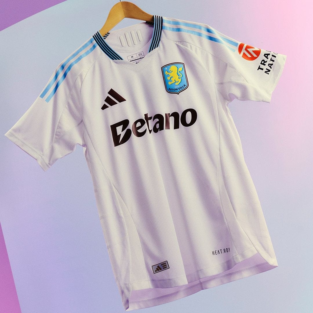 C6gCbrlQzGArPux | UAE SPORT STORE White Aston Villa football jersey on a hanger, Betano sponsor logo centered, with blue Adidas stripes and crest, against a pastel backdrop.
