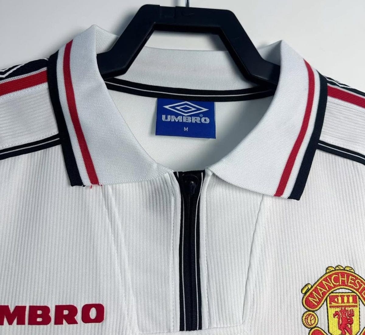 White Manchester United football jersey on a hanger, with blue Umbro tag and red/black collar trim, zipper at the front.