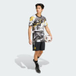 Male soccer player in a graphic black, white, and yellow jersey with black shorts, holding a soccer ball at his side.