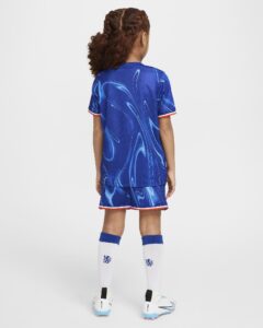 Back view of a young player in a blue abstract-pattern soccer jersey and shorts, white knee-high socks, and white cleats, standing against a white background with curly hair tied back.