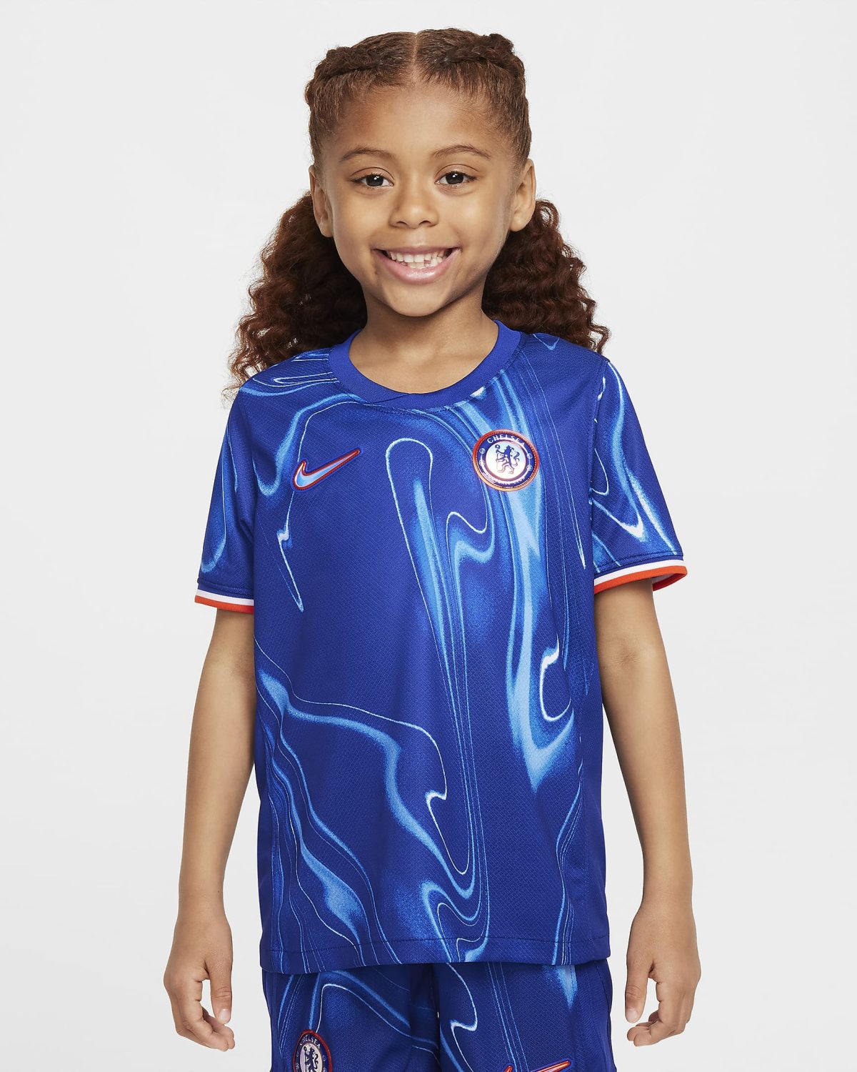 Smiling young girl wearing a blue Chelsea FC soccer jersey with crest, standing against a light background.