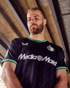 CNX0KQY8WUiSvIr | UAE SPORT STORE Portrait of a bearded man in a black soccer jersey with green trim standing under a concrete structure.