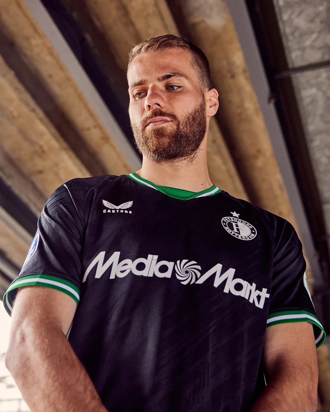 CNX0KQY8WUiSvIr | UAE SPORT STORE Portrait of a bearded man in a black soccer jersey with green trim standing under a concrete structure.