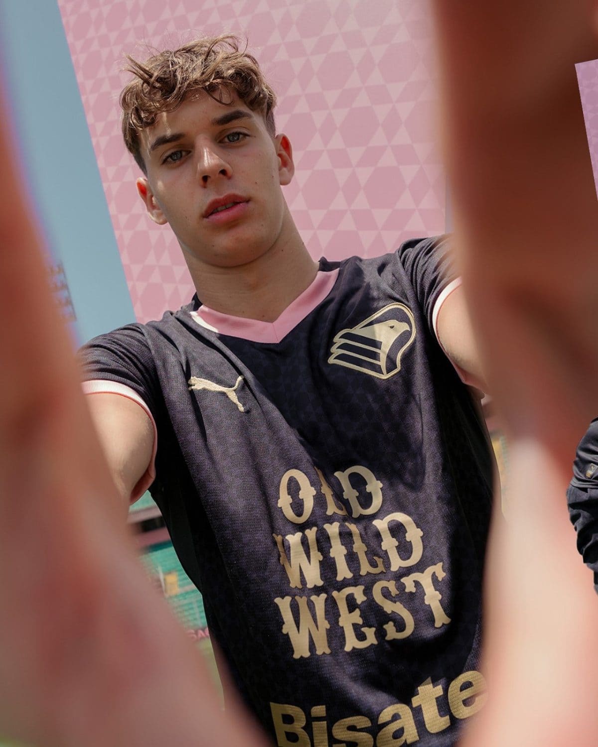 Close-up selfie of a young man looking at the camera, with hands in the foreground framing the shot. He wears a black jersey with pink collar and gold logos, against a pink geometric background.