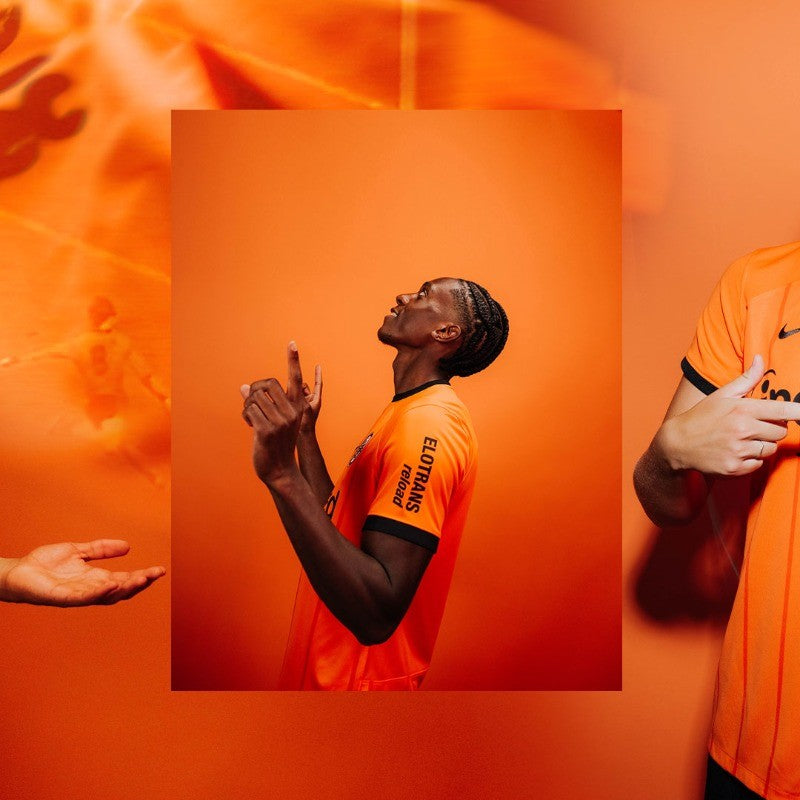 CSV0V9isaqJp2Lr | UAE SPORT STORE Portrait of a man in an orange jersey, looking up with hands raised, against an orange backdrop.