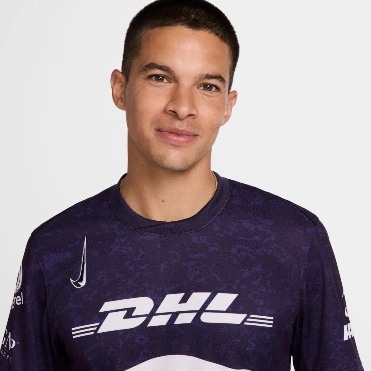 Portrait of a smiling man with short dark hair wearing a purple DHL jersey, facing the camera against a light gray background.