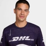 Portrait of a smiling man with short dark hair wearing a purple DHL jersey, facing the camera against a light gray background.