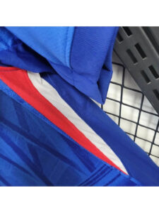 Camiseta-Chelsea-FC-1_-Equipacion-2526camisetafutboles_2-1080x1440-1 | UAE SPORT STORE Close-up of a blue jacket with a red and white stripe, resting on a black grid surface.