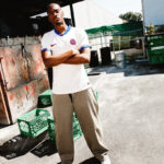 Man standing with arms crossed in a white PSG jersey, outdoors beside green crates and industrial structures.