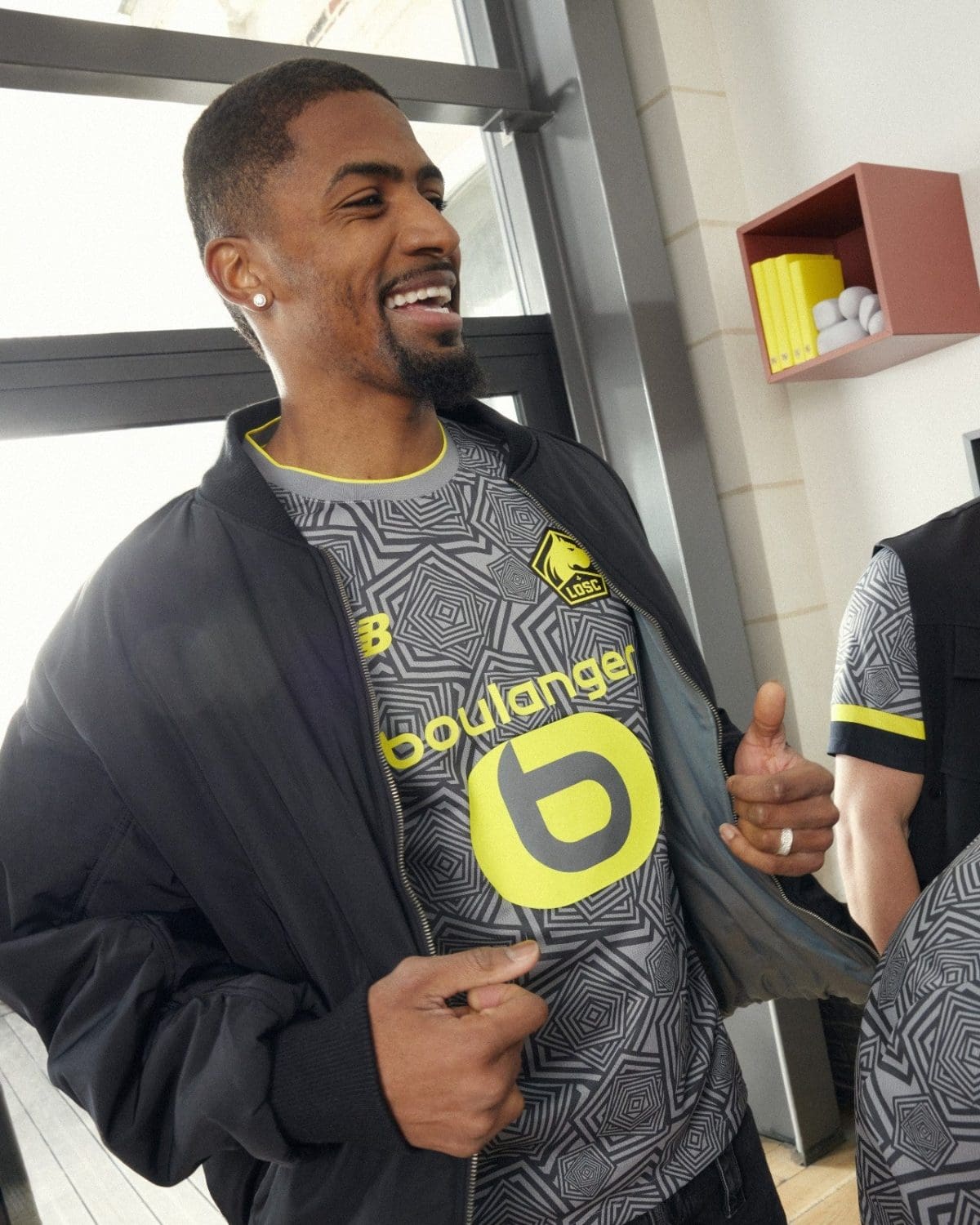Smiling man in a gray geometric training shirt and dark jacket, giving a thumbs-up indoors near a wall shelf with yellow books visible in the background.