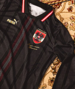 Black striped Puma sports jersey with crest badge and gold cursive text on a patterned rug backdrop.