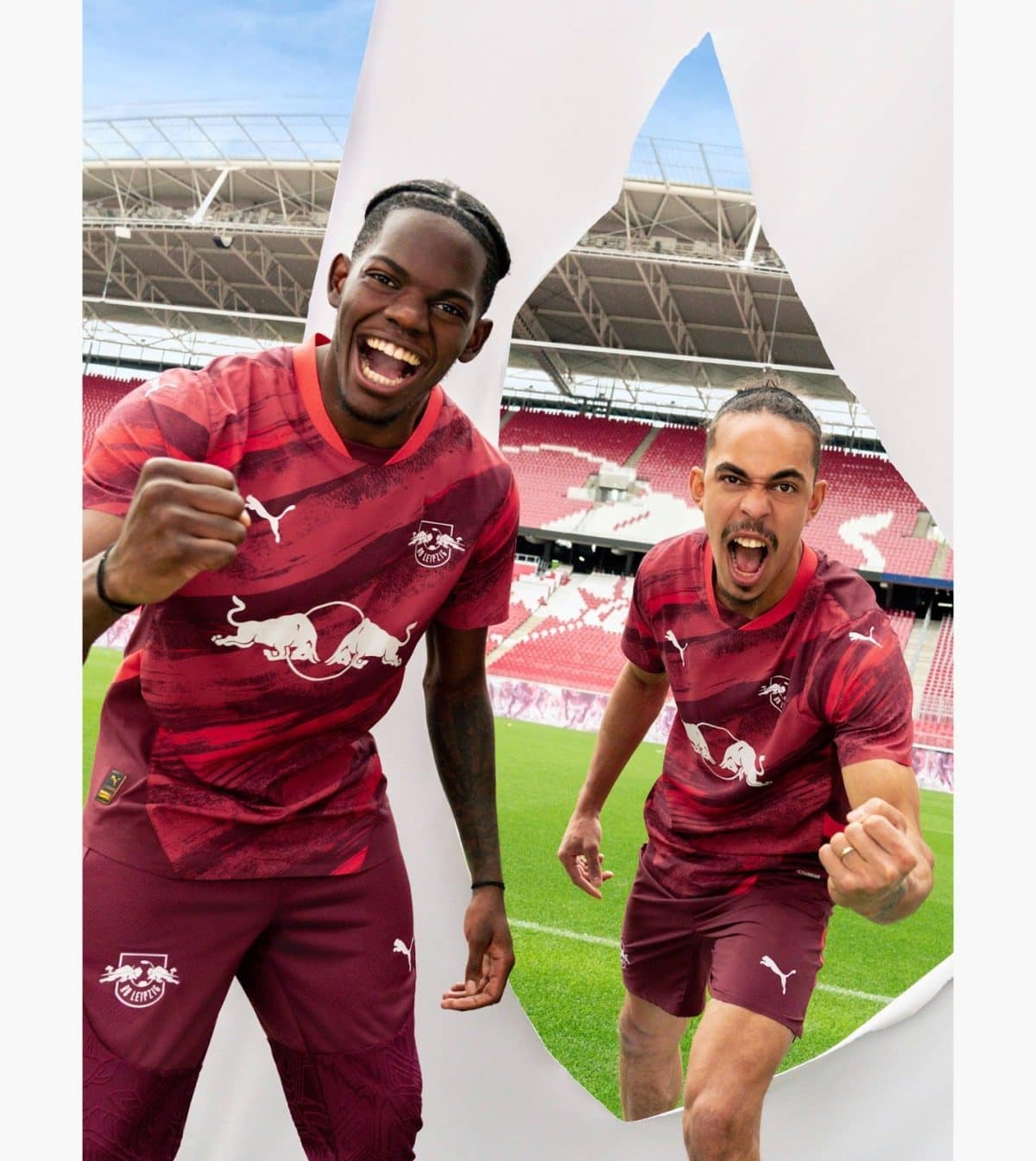 Two soccer players in maroon kits celebrate with clenched fists on the pitch, emerging from a torn white banner at a stadium.