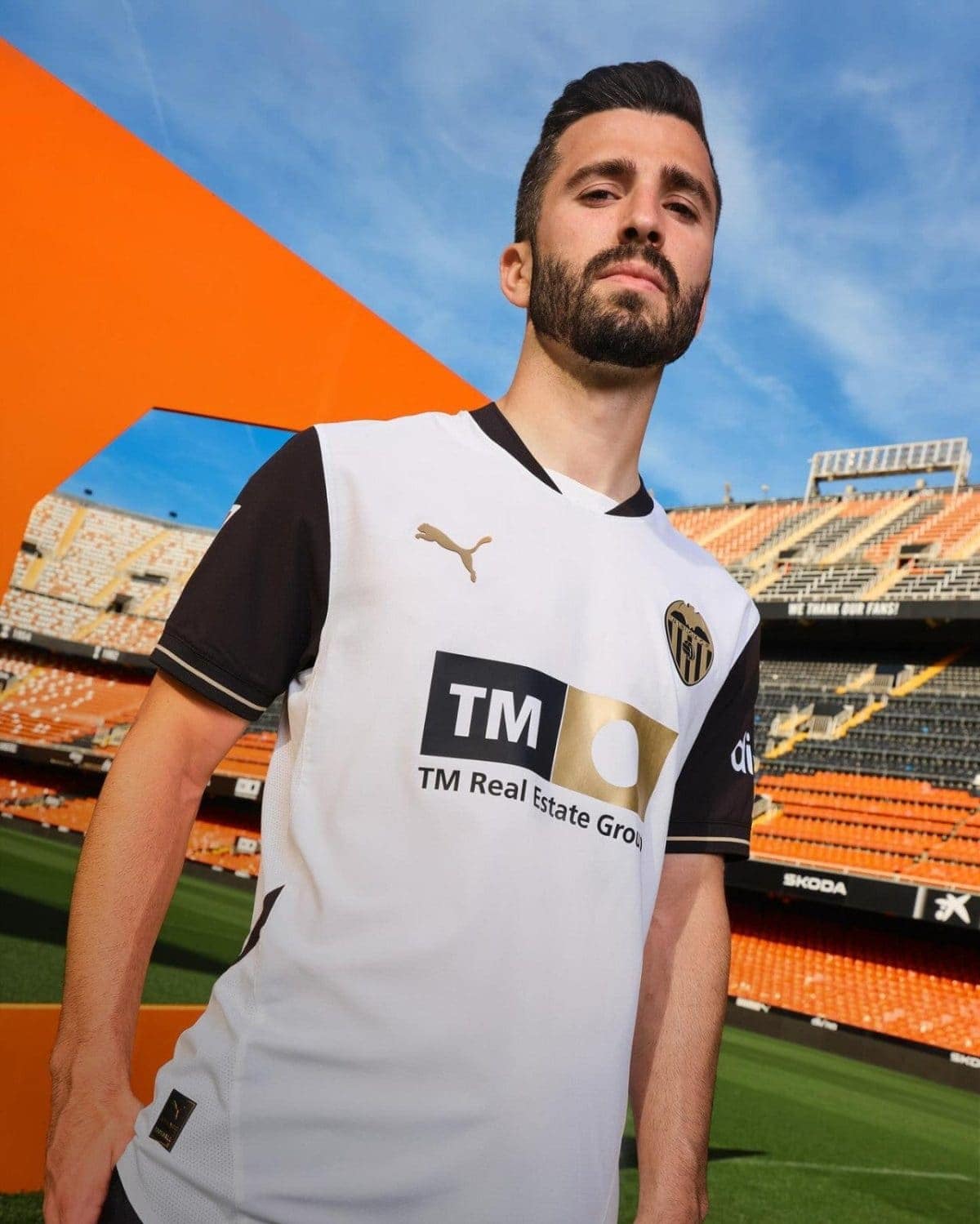 Male soccer player in Valencia CF jersey standing in an empty stadium with orange seats and blue sky behind him.