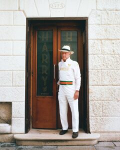 Older man in a white Venezia outfit with a fedora standing in a doorway of a stone building.
