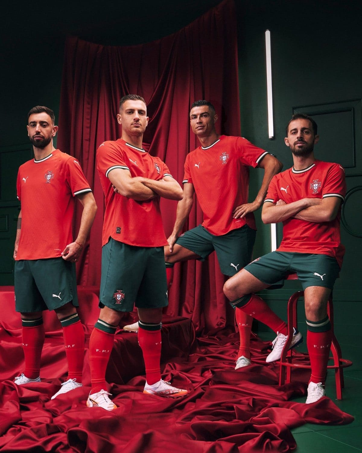 Four male Portuguese footballers pose in red Portugal jerseys and green shorts in a studio with red drapery, arms crossed or on hip, looking at camera.