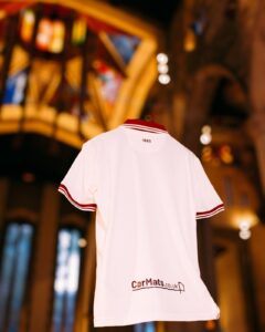 Back view of a white polo shirt with red striped collar and sleeve cuffs; '1889' at the neck and 'CarMats.co.uk' logo near the hem, blurred indoor background