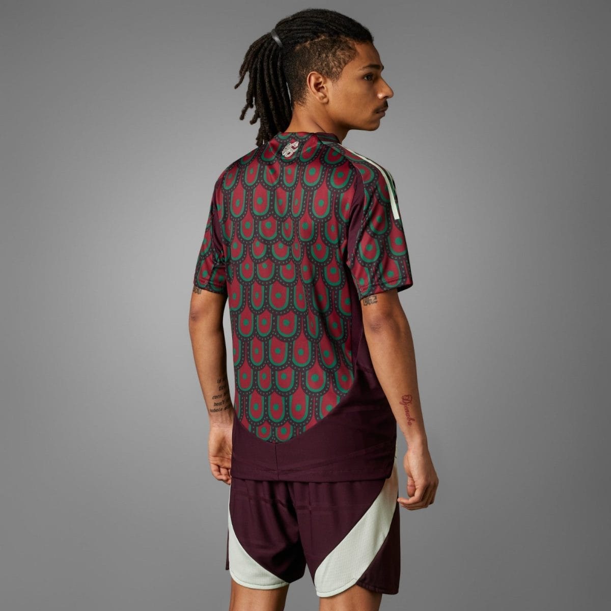 Person wearing a maroon and teal patterned sports jersey, viewed from the back against a gray studio backdrop with dreadlocks tied up.