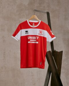 Red and white striped soccer jersey on a wooden hanger, featuring the UNIBET sponsor and a team crest on the chest, hanging against a beige wall.