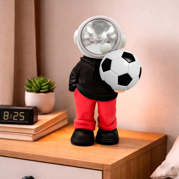Toy astronaut figure in a black hoodie and red pants stands on a wooden dresser, holding a soccer ball in front of a clock and plant.