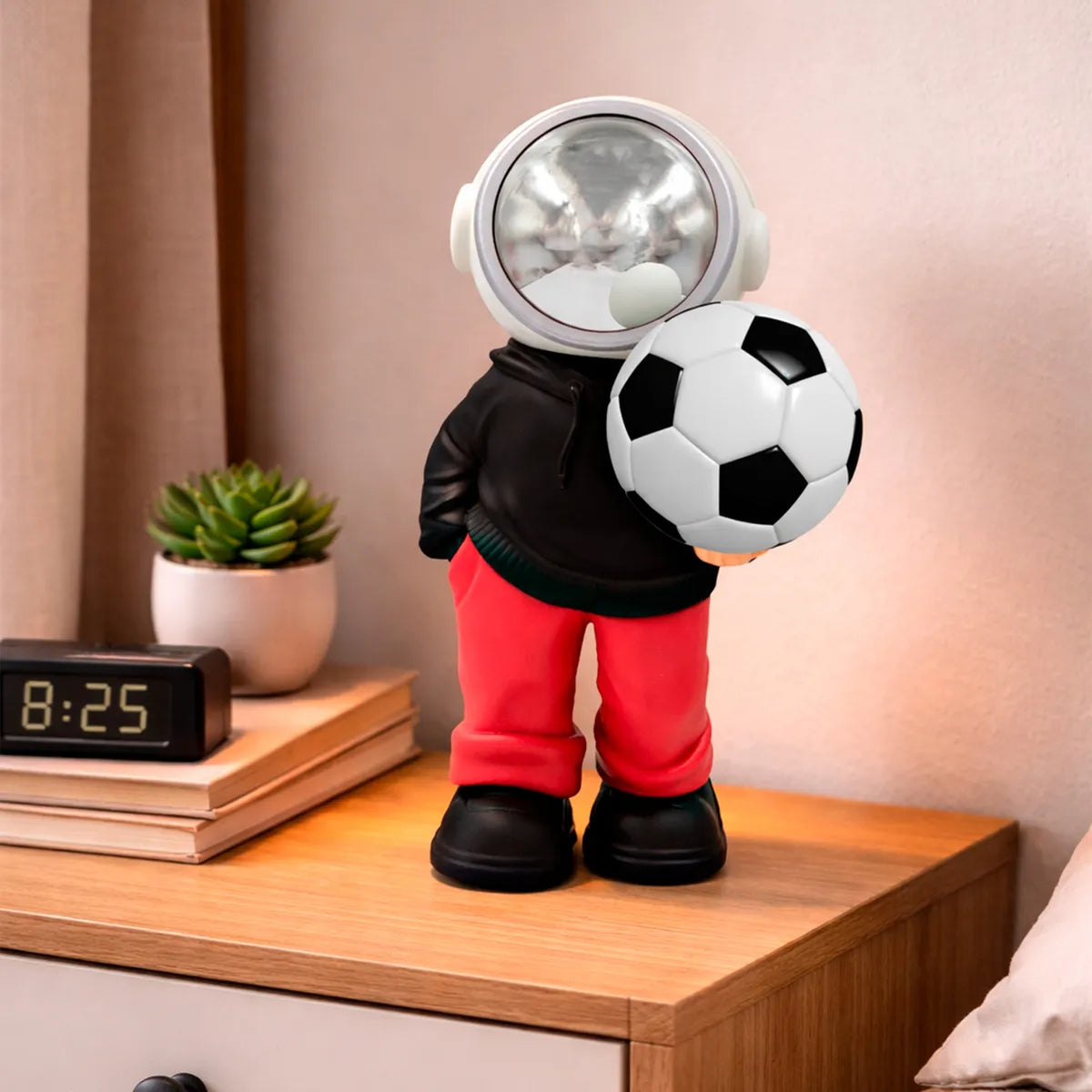 D_NQ_NP_2X_710685-MLM105767335041_012026-F-bocina-lampara-led-inalambrica-astronauta-balon-futbol | UAE SPORT STORE Toy astronaut figure in a black hoodie and red pants stands on a wooden dresser, holding a soccer ball in front of a clock and plant.