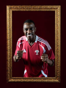 Smiling man in a red soccer jersey with fists up, standing inside a large ornate gold frame on a dark burgundy backdrop.