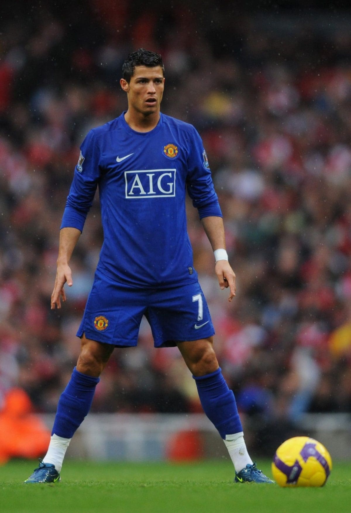 Manchester United player in a blue jersey stands on a green field in the rain with a yellow soccer ball nearby.